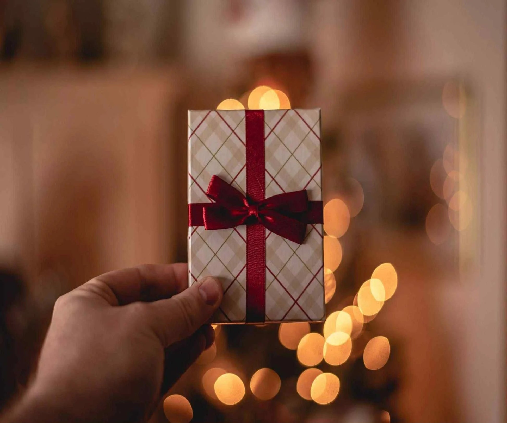 Hand holding a small gift box with a red ribbon against a blurred background with warm lights
