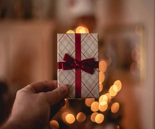 Hand holding a small gift box with a red ribbon against a blurred background with warm lights