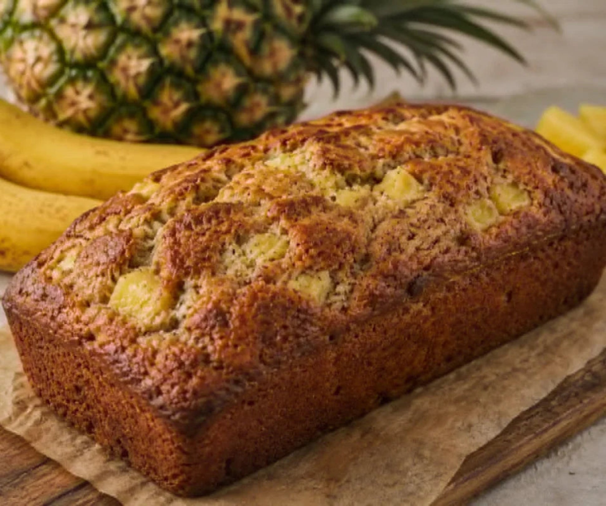 A loaf of pineapple bread on parchment paper with a whole pineapple and bananas blurred in the background.
