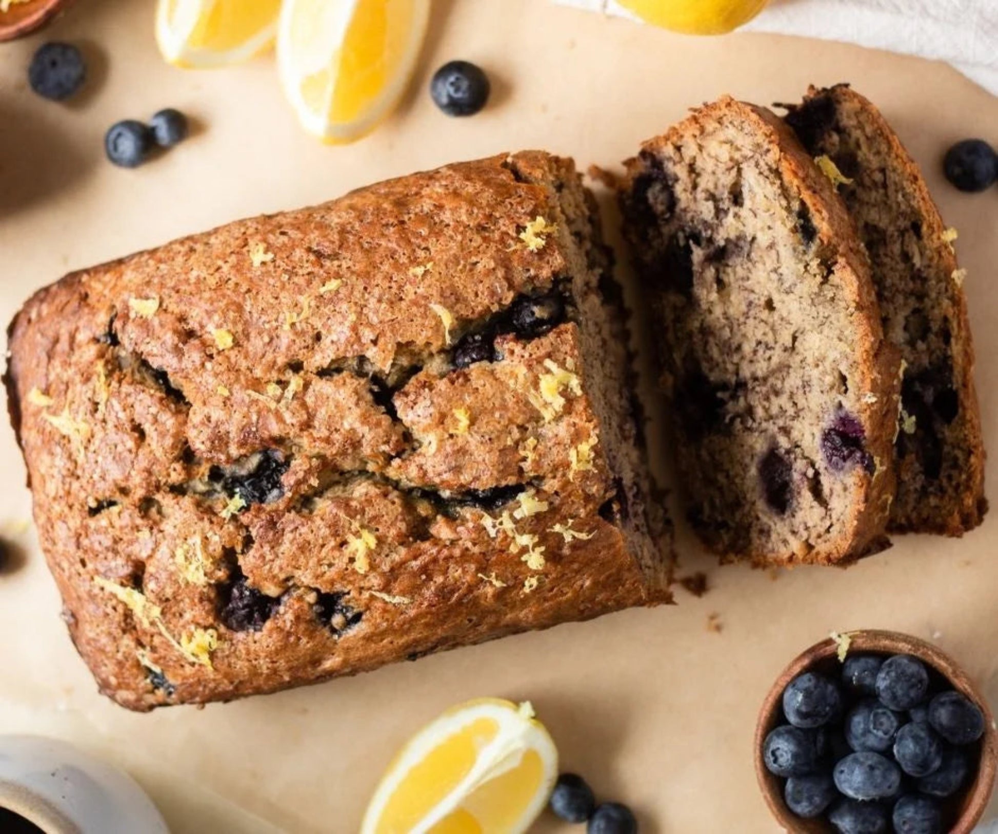 Loaf of blueberry bread with a slice cut, surrounded by blueberries and lemon wedges on a wooden surface.