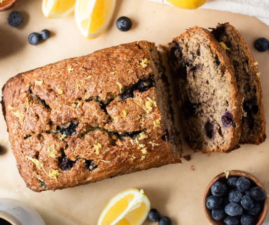 Loaf of blueberry bread with a slice cut, surrounded by blueberries and lemon wedges on a wooden surface.